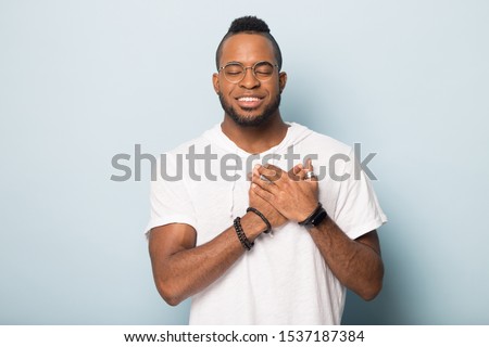 Smiling calm african American man in glasses isolated on blue studio background hold hands at heart chest praying, grateful biracial male believer feel happy peaceful, thanking god, faith concept