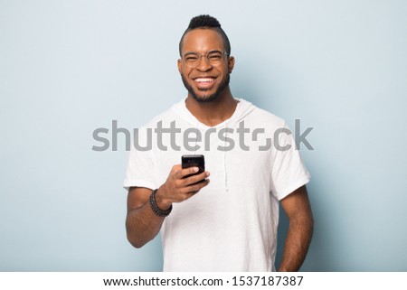 Overjoyed african American millennial man in casual wear and glasses isolated on blue studio background use modern cellphone look at camera, happy biracial male client excited with new smartphone app Royalty-Free Stock Photo #1537187387
