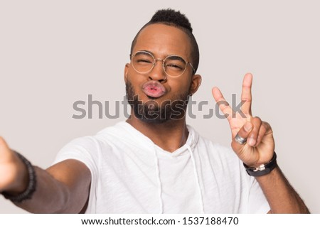 Funny biracial millennial male wear glasses isolated on grey studio background stretch hand posing for self-portrait picture, african American man make kiss taking selfie showing victory sign