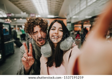 Cheerful emotional young couple posing together on camera. Taking selfie before flight. Travel together. Woman hold camera with hand. Guy show piece symbol. Trip of vacation honeymoon