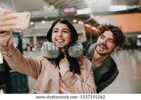 Cheerful happy attractive young couple stand together and take selfie. Woman hold smartphone in hand. She has travel pillow around neck. Guy stand behind her. Waiting for flight. Travel together