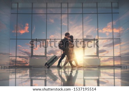 Picture of young couple walking in aurport close to window. Sunset outside. Going for a trip or vacation. Travel together. Take suitcase and backpacks
