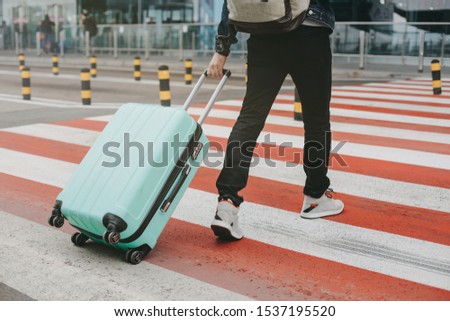 Picture of man walking on crosswalk and carrying blue suitcase with packpack on back. Going to airport or railway station alone. Cut view. Vacation or trip