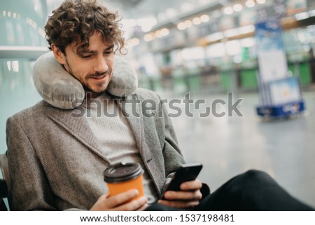 Picture of young man  sitting in waiting room and use phone in hands. Wait for train or airplane. Hold cup of coffee in hands. Pillow around neck. Calm and concentrated. Expert or manager travel abro