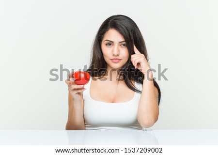 Young hispanic woman holding a tomato pointing his temple with finger, thinking, focused on a task.