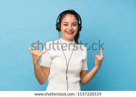 Young brunette girl over isolated blue background using the mobile with headphones and dancing