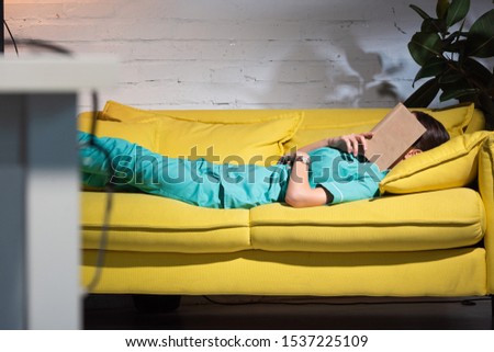 selective focus of nurse in uniform lying on sofa with book on face during night shift