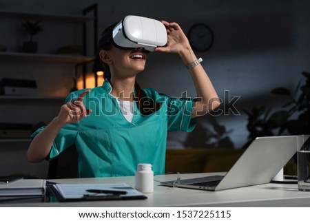 nurse in uniform with virtual reality headset pointing with finger during night shift Royalty-Free Stock Photo #1537225115