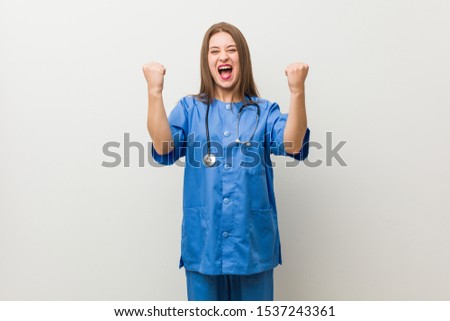 Young nurse woman against a white wall cheering carefree and excited. Victory concept.