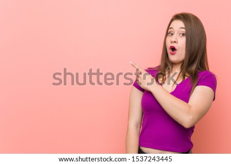 Young hispanic woman against a pink wall pointing to the side
