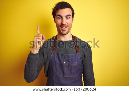 Young handsome shopkeeper man wearing apron standing over isolated yellow background pointing finger up with successful idea. Exited and happy. Number one.