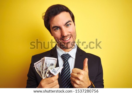 Young handsome businessman wearing suit holding dollars over isolated yellow background happy with big smile doing ok sign, thumb up with fingers, excellent sign