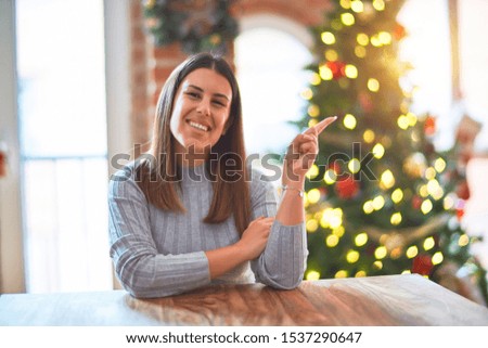 Young beautiful woman sitting at the table at home around christmas tree and decoration with a big smile on face, pointing with hand and finger to the side looking at the camera.