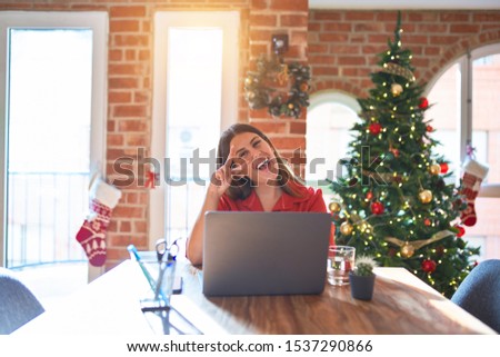 Beautiful woman sitting at the table working with laptop at home around christmas tree Doing peace symbol with fingers over face, smiling cheerful showing victory