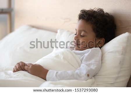 Peaceful cute little biracial boy sleeping in white cozy bed at home, happy smiling mixed race ethnicity preschooler child fall asleep relax in comfortable bedroom, having day rest daydreaming at home
