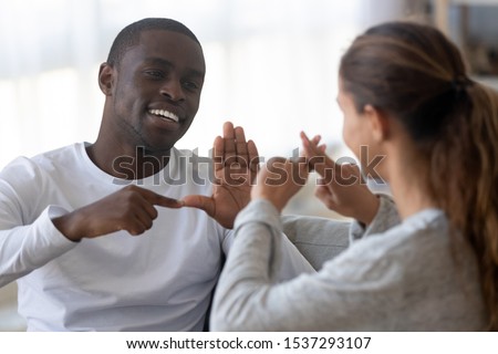 Smiling african American man sit on couch show hand gestures talking with female friend at home, international disabled hearing impaired couple or spouses use sign language communicating Royalty-Free Stock Photo #1537293107