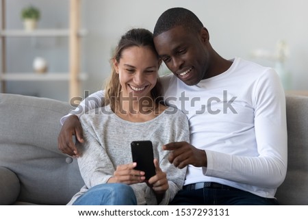 Happy multiethnic couple sit on couch at home watching funny video on smartphone, smiling loving international family husband and wife relax on sofa in living room using cellphone together Royalty-Free Stock Photo #1537293131