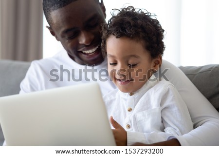 Happy loving african American young dad spend time playing on computer with little biracial son, smiling father relax at home with mixed race small boy child using laptop watch cartoons together