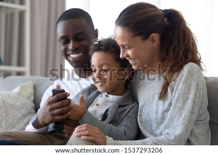 Happy international young family with little son sit on couch watching cartoons on cellphone together, smiling multiracial parents relax on couch at home with biracial boy child using smartphone