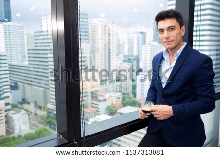 Adult handsome businessman who wears a formal suit is standing relaxed during a break from work, complete with a cup of coffee in a modern office surrounded by glass and have high-rise building views.