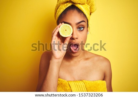 Afro woman wearing towel after shower holding slice lemon over isolated yellow background scared in shock with a surprise face, afraid and excited with fear expression