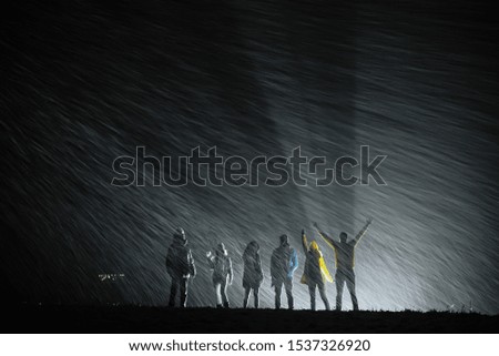Six happy friends are standing in back light and enjoying first snowfall at night time