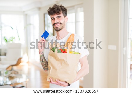 Young handsome man holding a paper bag full of fresh groceries at home, showing credit card as payment metod