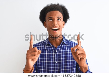Young african american man wearing casual shirt standing over isolated white background amazed and surprised looking up and pointing with fingers and raised arms.