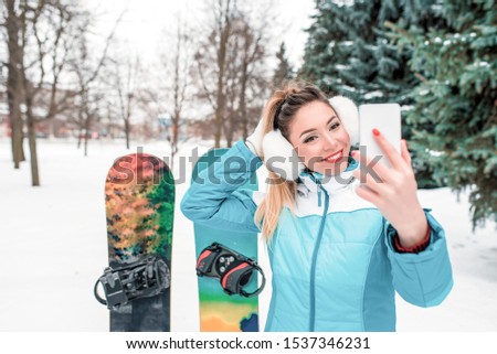 Beautiful woman, mobile phone talking video call, pictures selfie. Sports jumpsuit. Snowboards background snow, Christmas trees. Emotions happiness fun smile enjoyment winter resort. Free space text