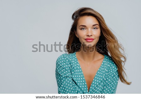 Studio shot of positive young woman has long wavy hair, makeup, wears turquoise polkadot blouse, looks straightly at camera, models against white background, copy space for your advertisement