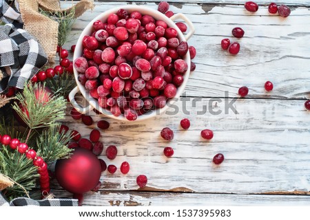 Frozen cranberries in a bowl over a white wood table background with Christmas decorations. Top view.