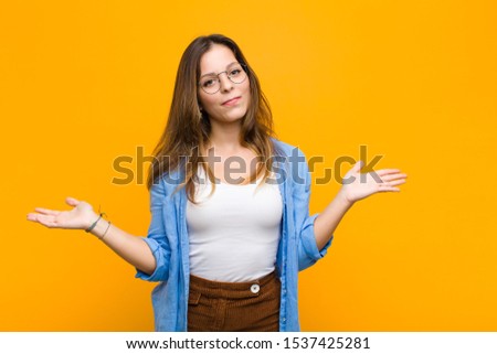 young pretty woman feeling puzzled and confused, unsure about the correct answer or decision, trying to make a choice against orange wall