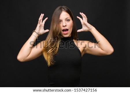 young pretty woman screaming with hands up in the air, feeling furious, frustrated, stressed and upset against black wall