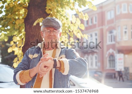 Stylish senior in fashionable clothes and in glasses stands in the city and cheks time on his watch.