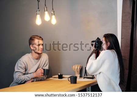 Young female photographer photographs a male model in a home setting by the light of warm lamps.