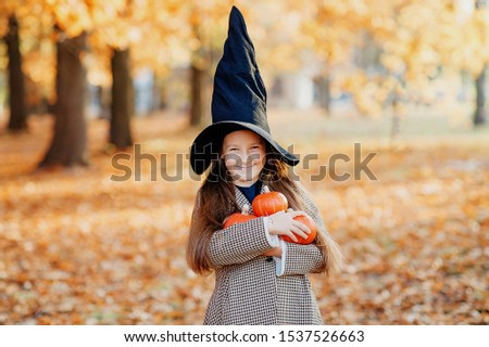 Little cute girl in the black witch hat with pumpkins Halloween decorations in the yellow autumn forest   