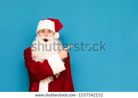 Portrait of surprised young Santa Claus, isolated on blue background, pointing finger at copyspace, looking into camera with surprised face. Shocked young Santa is showing up to empty space