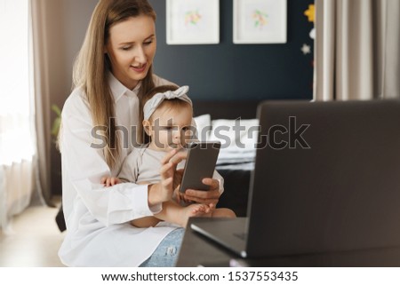Mother with baby daughter sitting in front of laptop and watching videos and photos on smartphone. Entertaining content for children on mobile phones, educating games, interesting cartoons, learning.
