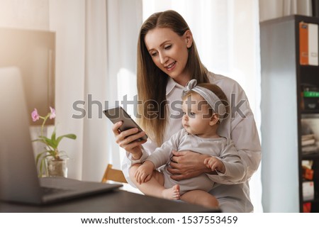 Little girl sitting on mother's knees and looking at screen of mom's smartphone. Young woman and daughter making video call in messenger app on mobile phone. Girl texting messages and comments in blog