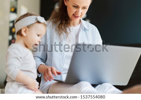 Young mother doing online job while sitting at home with child. Mom entertaining little kid with educating cartoons and interactive Internet channels for children. Woman making video call on laptop.