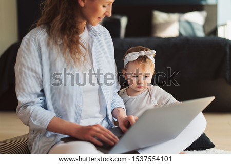 Mother showing interesting cartoons on laptop to daughter. Young woman and baby girl sitting on floor and looking at screen of wireless device.Mom doing work online remotely at home,child sitting near