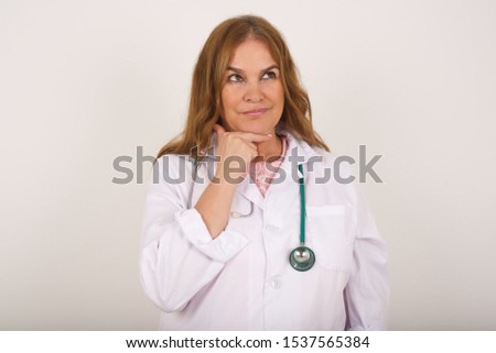 Dreamy doctor female with pleasant expression, wearing medical uniform, looks sideways, keeps hand under chin, thinks about something pleasant, poses against gray background.