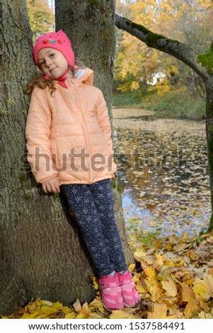 A five-year-old girl stands leaning to a tree on the bank of a pond in an autumn park