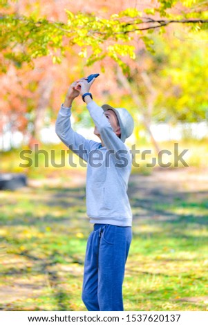 A boy in a hat photographs autumn leaves at the top on a smartphone in the park.