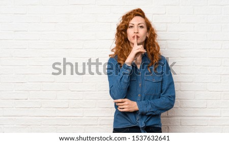 Redhead woman over white brick wall showing a sign of silence gesture putting finger in mouth