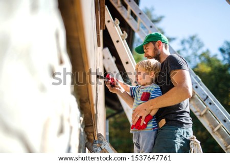 A father and toddler boy outdoors in summer, painting wooden house.