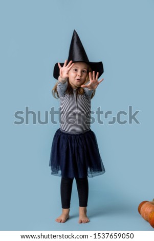 Little girl in a witch costume standing at the blue studio, pulls hands and shows fingers. Studio shot