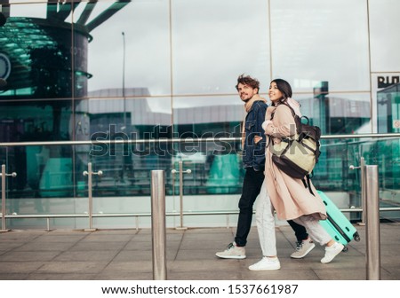 Picture of young couple walking together close to airport. Ready to travel. Carry suitcase or backpack. In rush. Trip or honeymoon vacation. Walk in front of glass wall