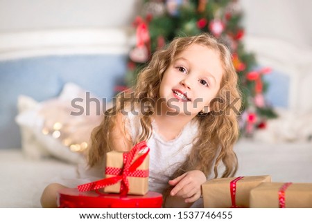 Portrait of a little emotional girl on the bed on the background of Christmas decorations. Christmas tree in the background. Gifts with red ribbons in her hands.