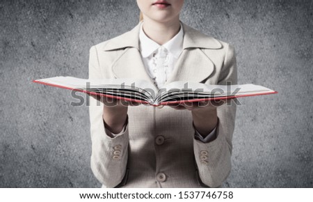Business woman holding open book on background of grey wall. Business knowledges and education concept. Closeup open book with red cover in female hands. Elegant young woman in jacket.